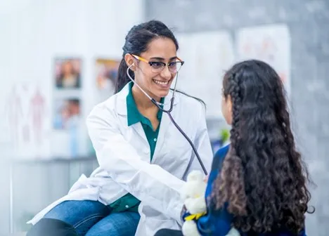 A nurse practitioner in a white coat listens to a young patient’s heart with a stethoscope